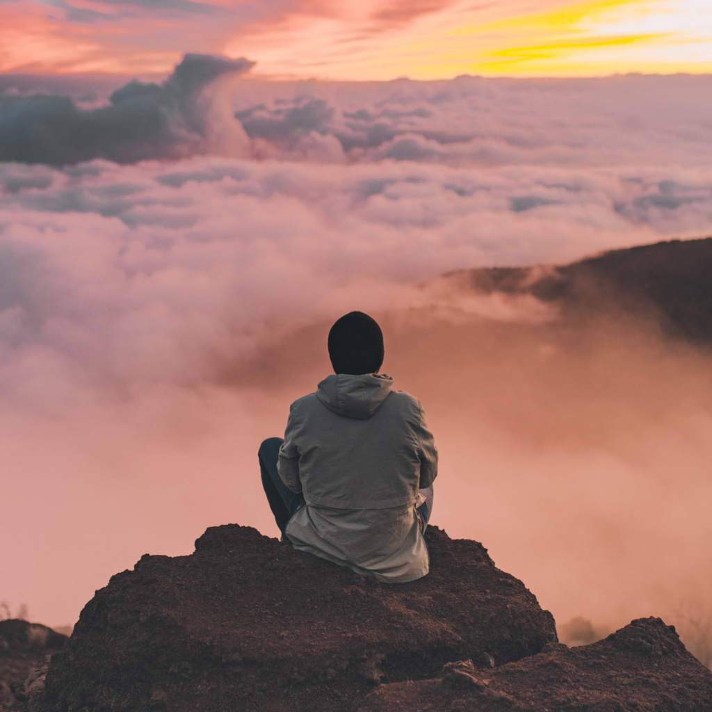 man sitting on cliff clouds pink sunrise