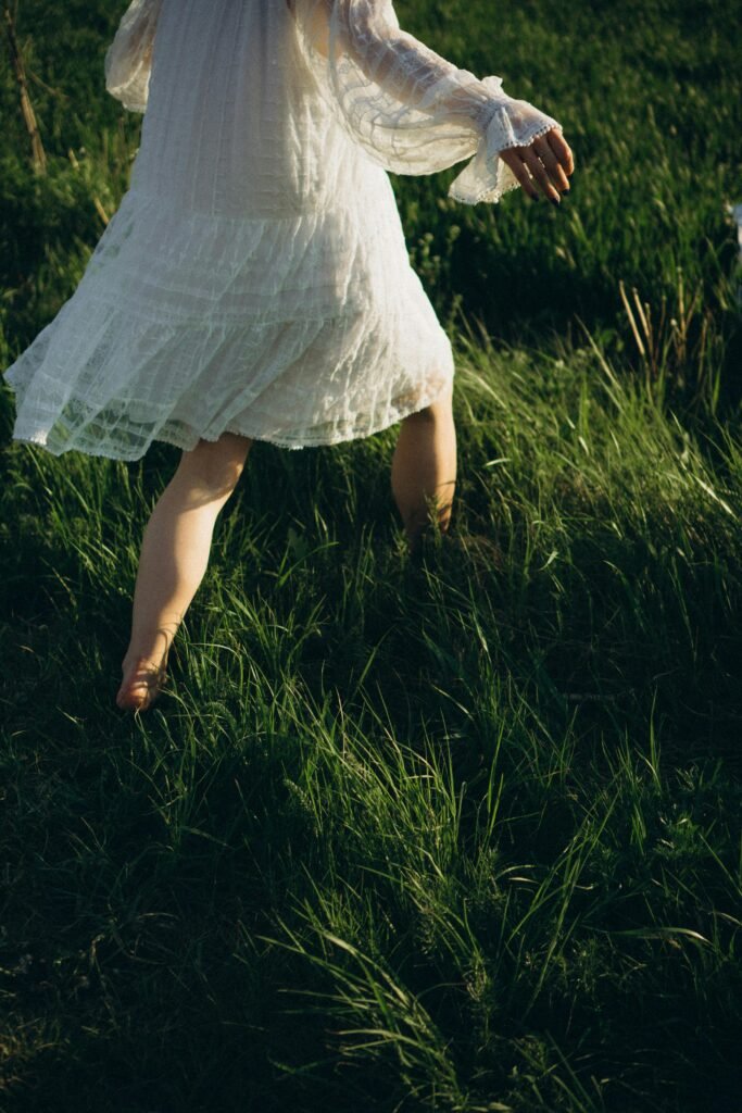 A woman in a white lace dress walking through a sunlit grassy field.