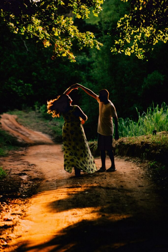 A couple gracefully dances on a sunlit dirt road surrounded by lush greenery in Saquarema, Brazil.