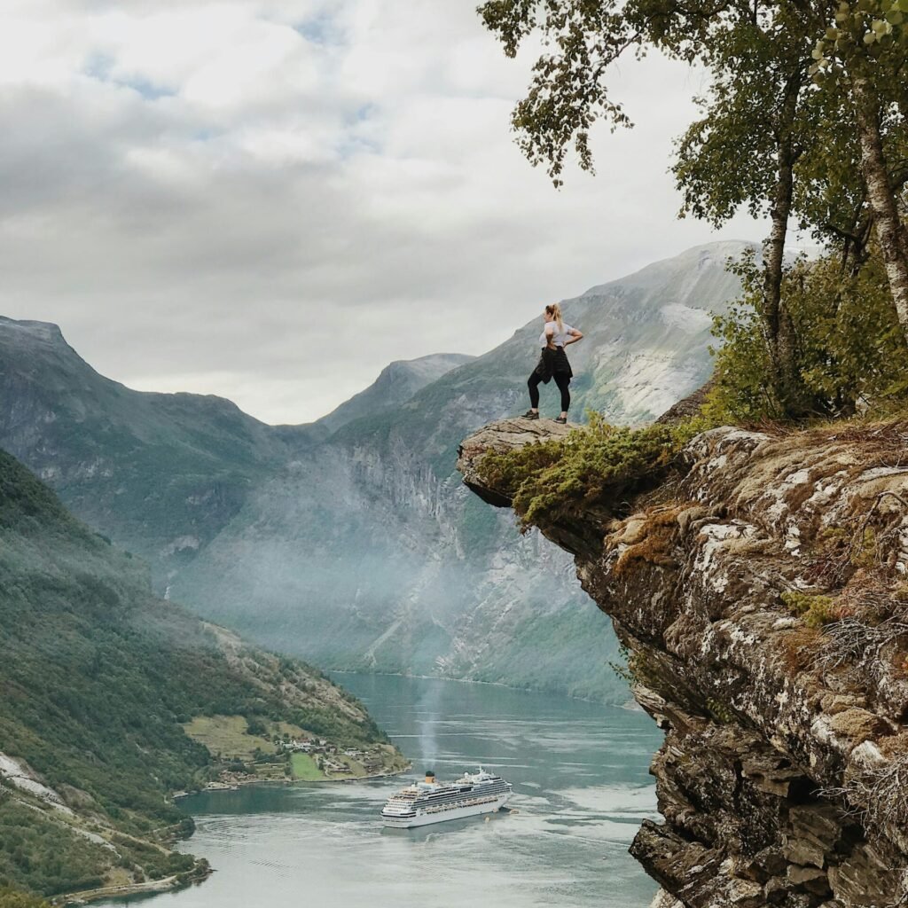 Person enjoys breathtaking view from a cliff above Geiranger Fjord, Norway, with clear skies and cruise ship below.