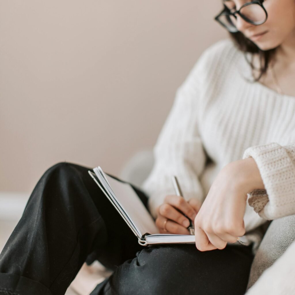 Attentive female wearing eyeglasses and casual outfit sitting barefoot with crossed legs on comfortable couch in modern flat and taking notes in notebook with pen