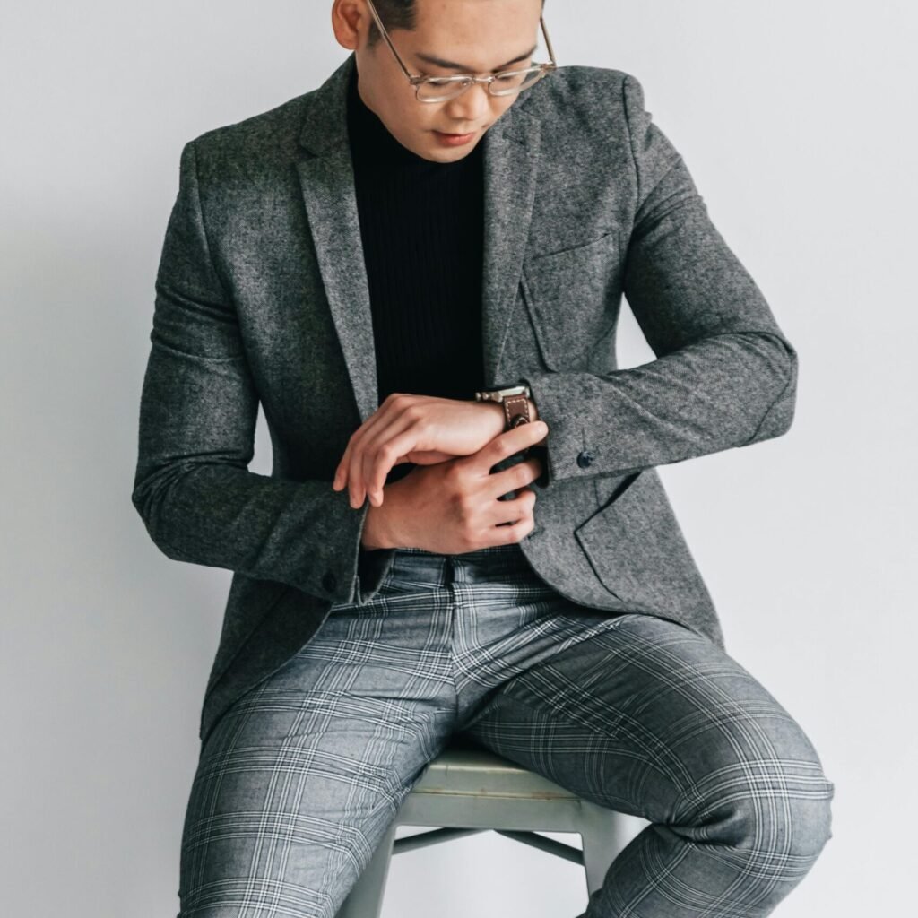 A fashionable young man in a gray suit is seated while checking his watch.