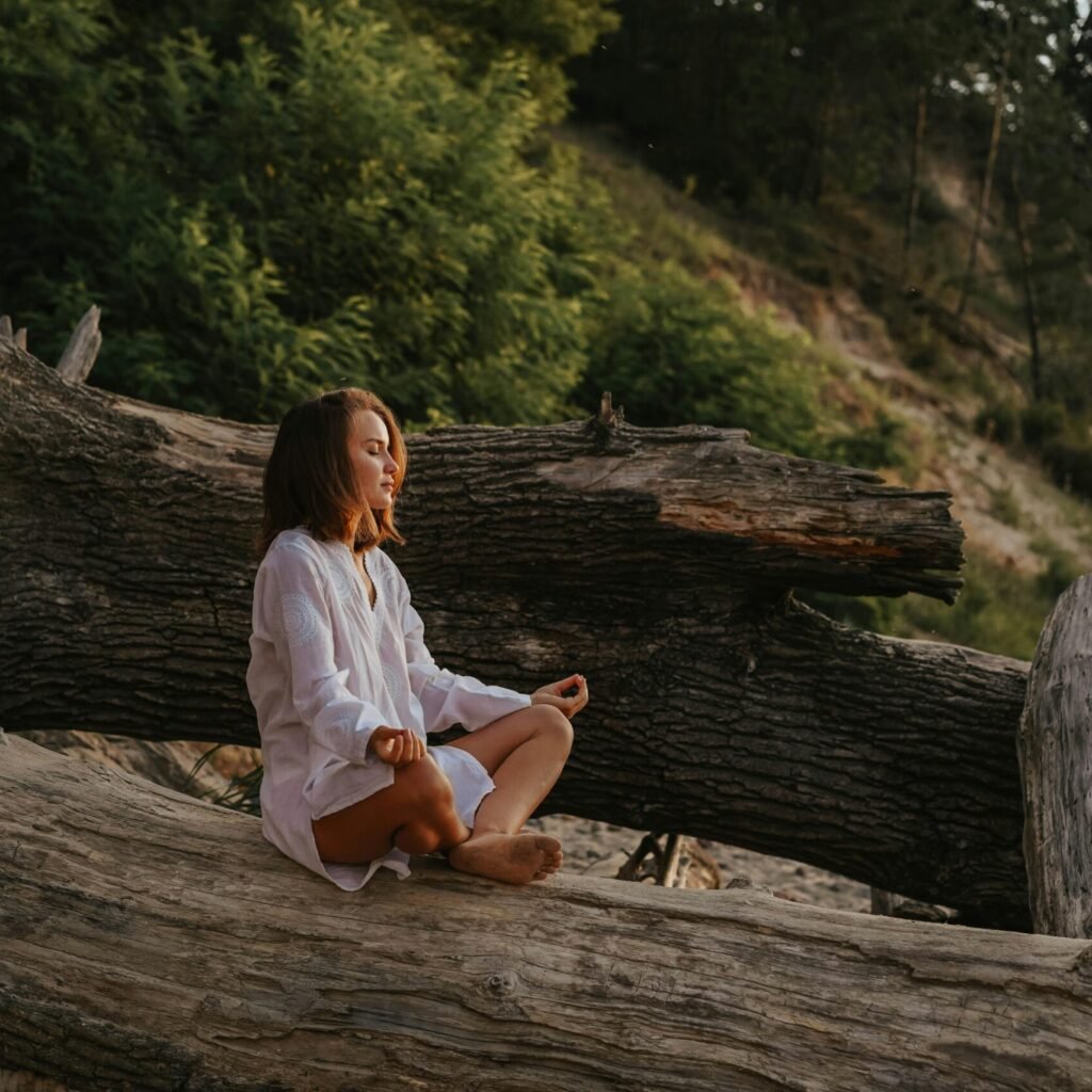 Woman in white shirt meditates on a large log in a forest setting during the day.