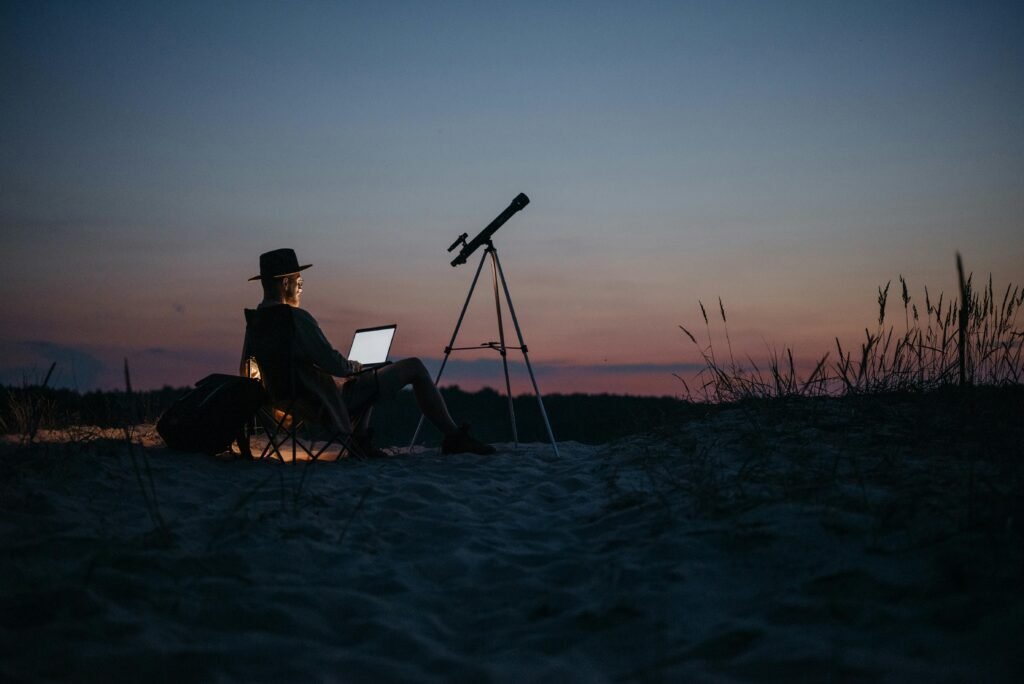 Person using a laptop and telescope at twilight, combining work and stargazing in a serene outdoor setting.