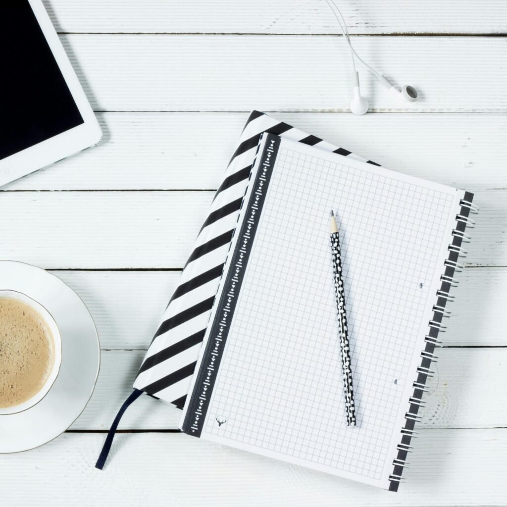 Flatlay of a modern workspace featuring coffee, tablet, and notebook with pencil on a white wooden desk.